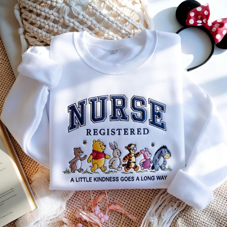 May include: White sweatshirt with the word "NURSE" in navy blue collegiate lettering, with "REGISTERED" below. Features a line of cartoon characters and the text "A LITTLE KINDNESS GOES A LONG WAY".