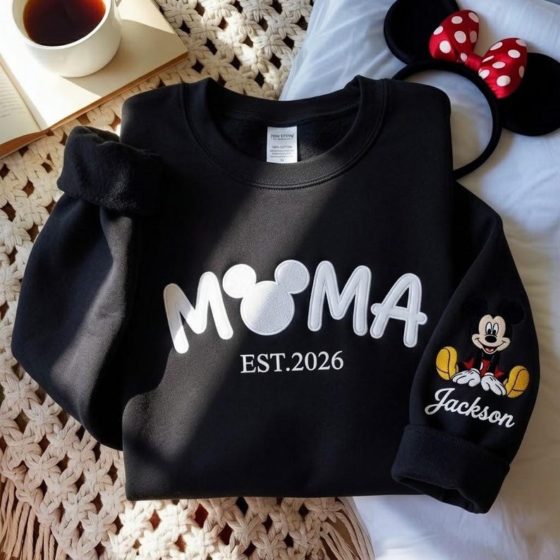 May include: Black sweatshirt with the word "M♡MA" in white, with a Mickey Mouse head replacing the "O". The sleeve features a Mickey Mouse graphic and the name "Jackson". The text "EST.2026" is below the word "M♡MA".