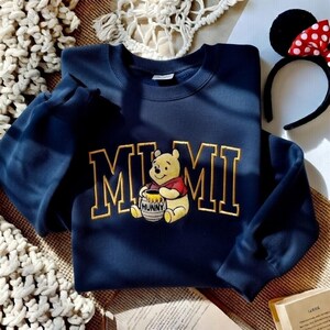 以下が含まれることがあります： 金色の文字で「MIMI」と書かれたネイビーブルーのスウェットシャツと、蜂蜜の壺を持ったプーさんの刺繍グラフィック。赤と白のドット柄のリボンが付いた黒いカチューシャが背景に見えます。
