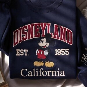 Puede incluir: Sudadera azul marino con un dise&ntilde;o de Disneyland. El dise&ntilde;o incluye la palabra "Disneyland" en granate y blanco, el a&ntilde;o "1955" y la palabra "California". Tambi&eacute;n hay un gr&aacute;fico de Mickey Mouse.