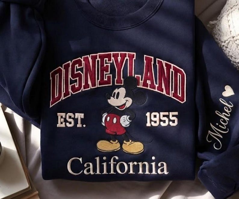 Puede incluir: Sudadera azul marino con un dise&ntilde;o de Disneyland. El dise&ntilde;o incluye la palabra "Disneyland" en granate y blanco, el a&ntilde;o "1955" y la palabra "California". Tambi&eacute;n hay un gr&aacute;fico de Mickey Mouse.