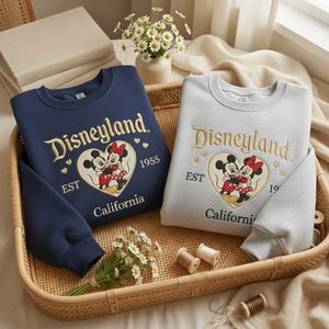 May include: Two sweatshirts, one navy blue and one light gray, featuring the Disneyland logo with Mickey and Minnie Mouse in a heart. The sweatshirts have long sleeves and are displayed in a woven basket with flowers.