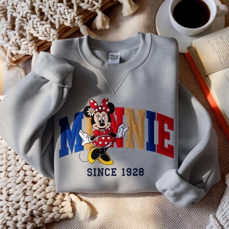 K&ouml;nnte beinhalten: Ein graues Sweatshirt mit einem farbenfrohen Minnie Mouse-Motiv. Das Design zeigt den Namen "MINNIE" in Blau, Gold und Rot, mit Minnie Mouse in einem roten gepunkteten Kleid. Der Text "SINCE 1928" steht darunter.