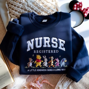 May include: Navy blue sweatshirt with the word "NURSE" in white, embroidered in a collegiate style. Below, the word "REGISTERED" is embroidered. A line of cartoon characters is below the text, with the phrase "A LITTLE KINDNESS GOES A LONG WAY".