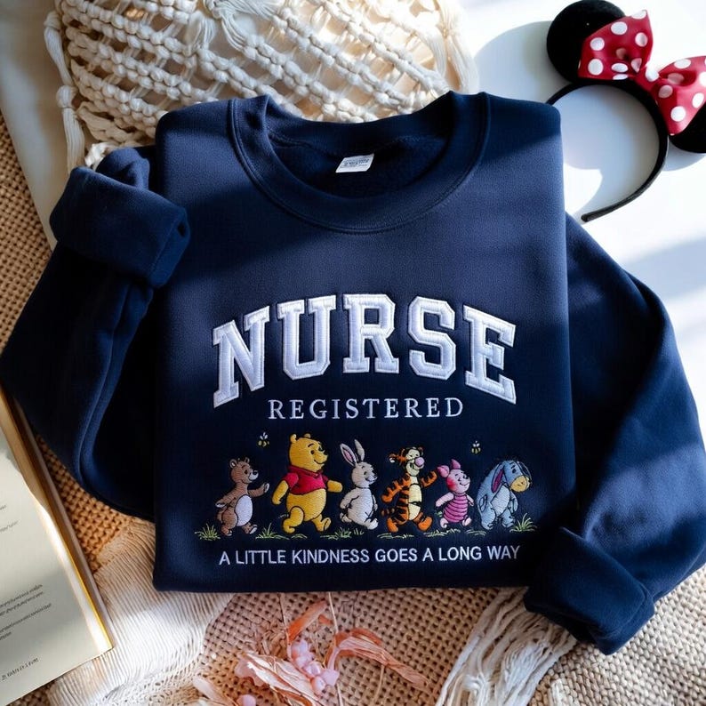 May include: Navy blue sweatshirt with the word "NURSE" in white, embroidered in a collegiate style. Below, the word "REGISTERED" is embroidered. A line of cartoon characters is below the text, with the phrase "A LITTLE KINDNESS GOES A LONG WAY".