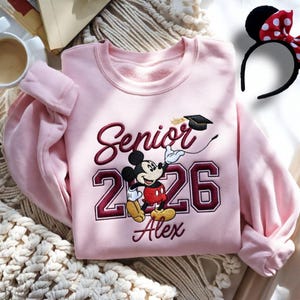 May include: A light pink sweatshirt with the embroidered words "Senior 2026" and a Mickey Mouse graphic wearing a graduation cap. The name "Alex" is embroidered below the numbers. A black headband with a red and white polka dot bow is also visible.