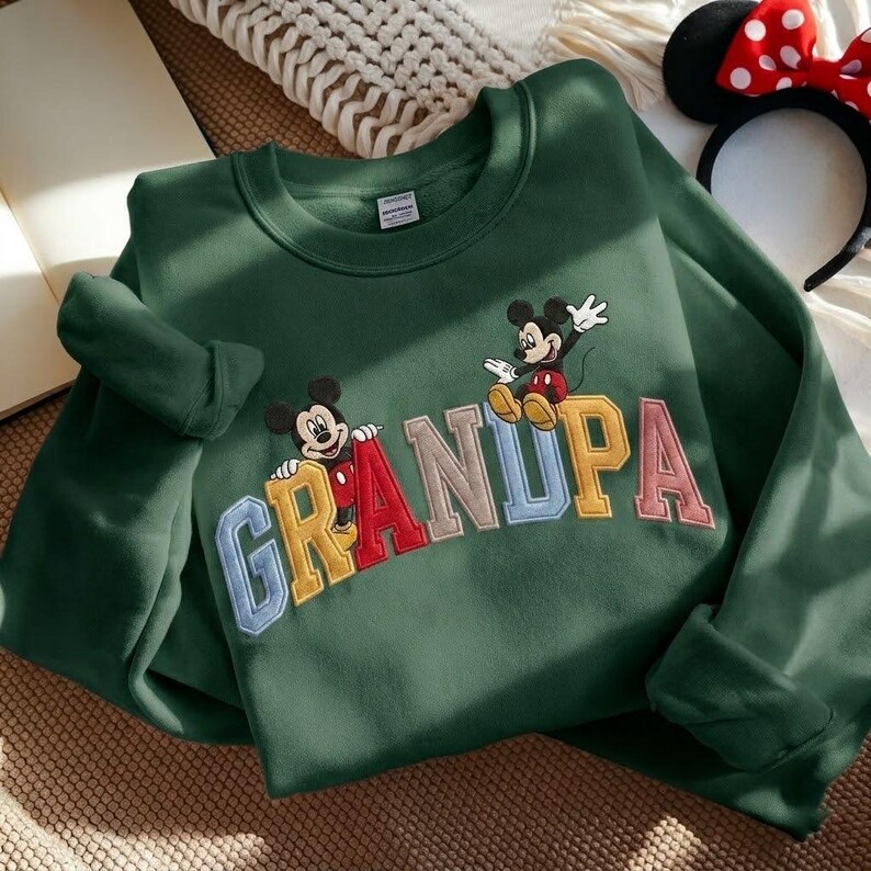 May include: A dark green sweatshirt with the word "GRANDPA" embroidered in colourful letters. Two Mickey Mouse figures are featured above the letters. A Minnie Mouse headband is visible in the background.