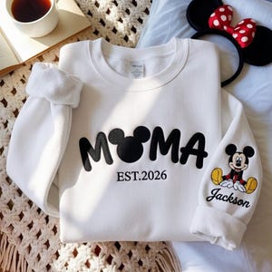 May include: White sweatshirt with the word "MOMA" in black, with a Mickey Mouse head replacing the "O". The text "EST.2026" is below. The right sleeve features a Mickey Mouse graphic and the name "Jackson". A Minnie Mouse headband is in the background.