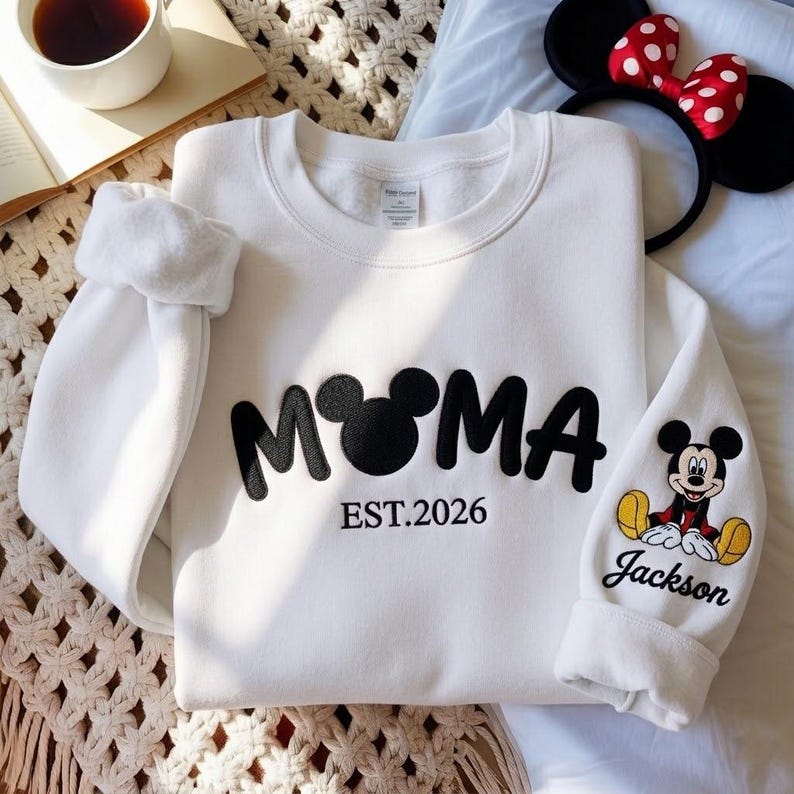 May include: White sweatshirt with the word "MOMA" in black, with a Mickey Mouse head replacing the "O". The text "EST.2026" is below. The right sleeve features a Mickey Mouse graphic and the name "Jackson". A Minnie Mouse headband is in the background.