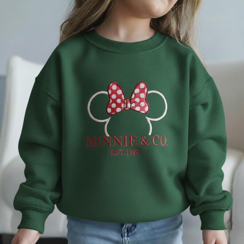 Op de afbeelding: Donkergroene sweatshirt met een Minnie Mouse-ontwerp. Het ontwerp bevat een rode strik met witte stippen, witte muizenoren en de tekst "MINNIE & CO. EST. 1928" in het rood. De sweatshirt is effen groen van kleur.
