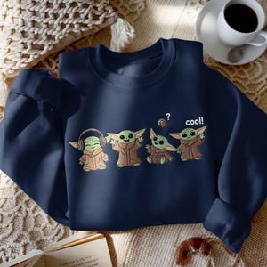 May include: Navy blue sweatshirt featuring a cartoon design of four Grogu characters in different poses. One Grogu wears headphones, and the others have various expressions. The word "cool!" is printed on the sweatshirt.