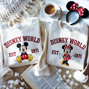 May include: Two cream-colored sweatshirts with the words "DISNEY WORLD EST 1971" embroidered in red. One features Mickey Mouse, the other Minnie Mouse. A black headband with red polka dot bow is visible.