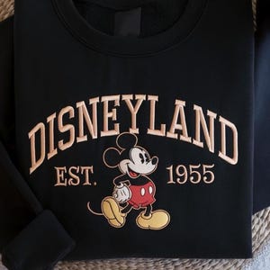 May include: Black sweatshirt featuring the word "DISNEYLAND" in tan embroidery. Below the word is "EST. 1955" and a cartoon image of Mickey Mouse in red shorts and yellow shoes.