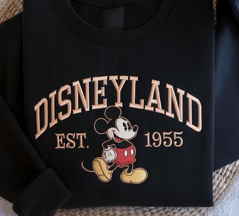 May include: Black sweatshirt featuring the word "DISNEYLAND" in tan embroidery. Below the word is "EST. 1955" and a cartoon image of Mickey Mouse in red shorts and yellow shoes.