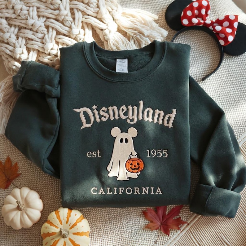 Pode incluir: Uma camisola de gola redonda verde-escura com o texto bordado Disneyland est 1955 California. O design central apresenta um fantasma branco com orelhas de rato a segurar um balde de ab&oacute;bora cor de laranja tipo lanterna.