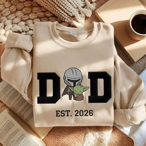 May include: A beige sweatshirt with the word "DAD" in large black block letters, featuring an embroidered image of a Mandalorian and Grogu. Below the text is "EST. 2026" in black. The sweatshirt has long sleeves and a crew neck.