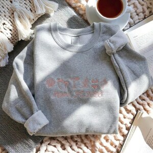 Peut inclure: Un sweat-shirt gris avec des manches retrouss&eacute;es, brod&eacute; de motifs roses sur le th&egrave;me de la plage et du texte "Miami, FL 2026". Le motif comprend un coquillage, des palmiers, un cocktail et un bikini.