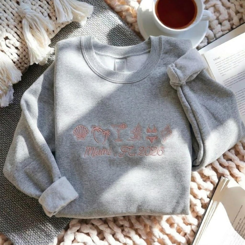 Peut inclure: Un sweat-shirt gris avec des manches retrouss&eacute;es, brod&eacute; de motifs roses sur le th&egrave;me de la plage et du texte "Miami, FL 2026". Le motif comprend un coquillage, des palmiers, un cocktail et un bikini.