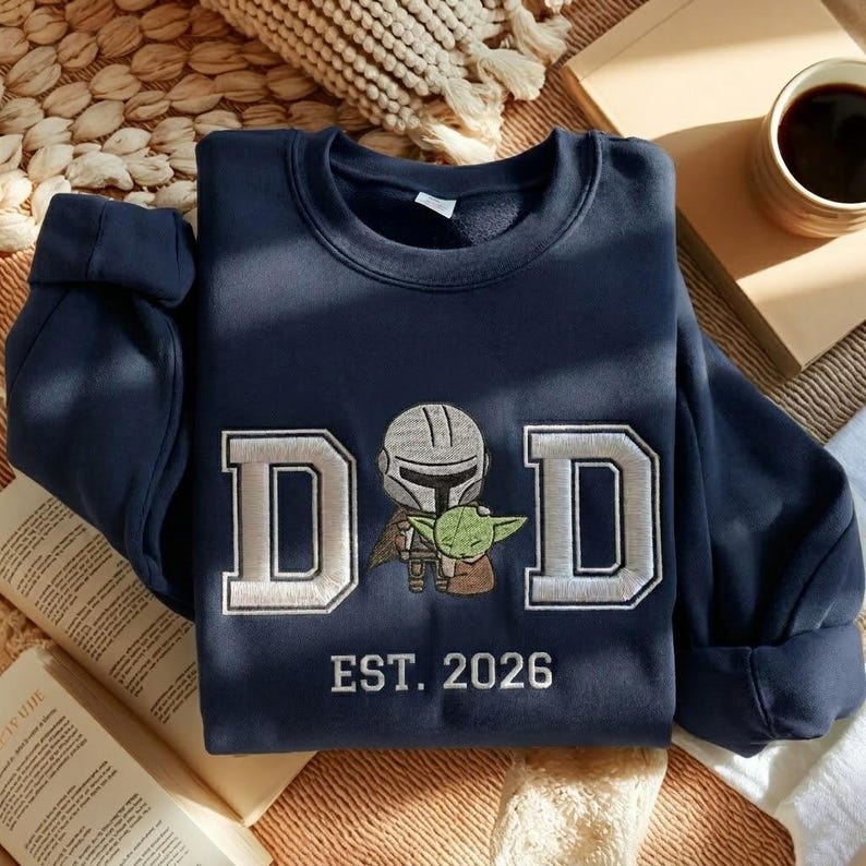 May include: Navy blue sweatshirt with the word "DAD" in white block letters. An embroidered image of a Mandalorian and Grogu is centered between the letters. The text "EST. 2026" is below the image.