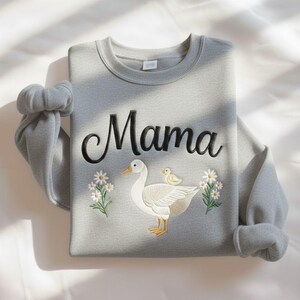 May include: A light gray sweatshirt with the word "Mama" embroidered in black script. Below the text is an embroidered design of a white goose with a yellow chick and white daisies. The sleeves are rolled up.