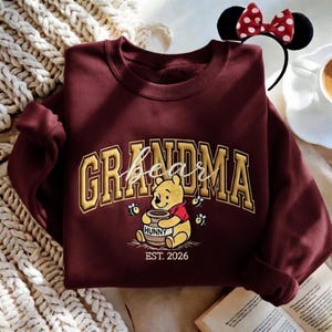 May include: Burgundy sweatshirt with the word "Grandma" in gold lettering, featuring Winnie the Pooh holding a honey pot. The sweatshirt also has the text "EST. 2026". A Minnie Mouse ear headband is in the background.