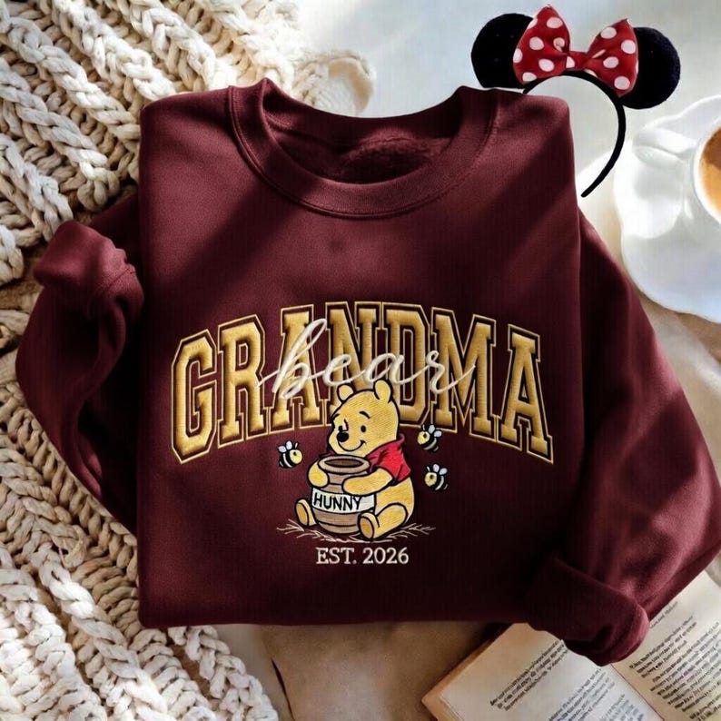 May include: Burgundy sweatshirt with the word "Grandma" in gold lettering, featuring Winnie the Pooh holding a honey pot. The sweatshirt also has the text "EST. 2026". A Minnie Mouse ear headband is in the background.