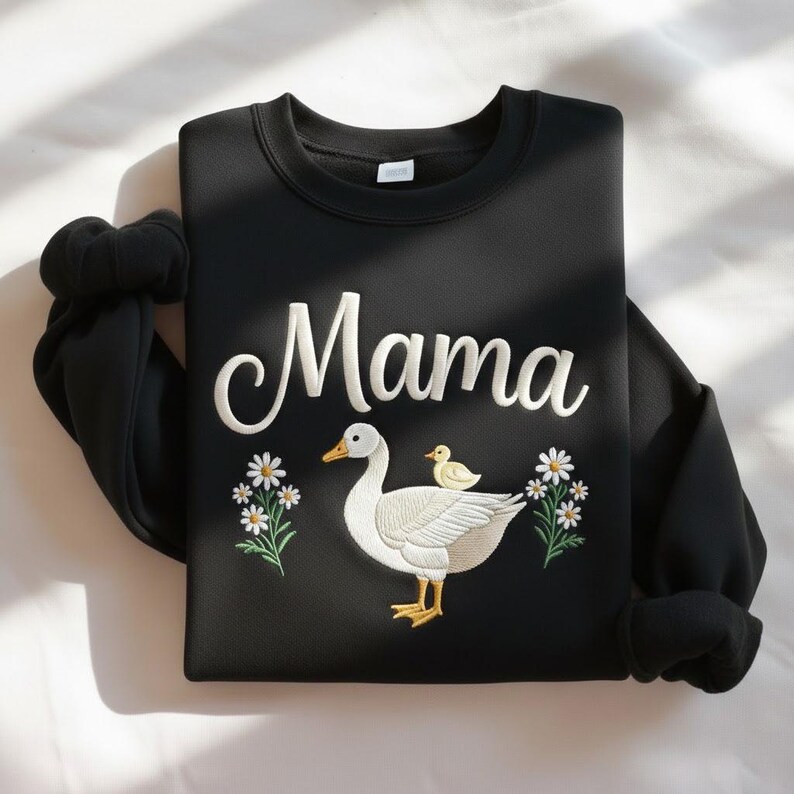 May include: Black sweatshirt with the word "Mama" in white script. The design features a white goose with a yellow beak and feet, and a yellow chick. White daisies with green stems and leaves are on either side of the goose.