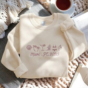 Peut inclure: Sweat-shirt cr&egrave;me avec broderie &laquo; Miami, FL 2026 &raquo;. Le motif comprend un coquillage, un palmier, un cocktail, une plante tropicale, un bikini et un autre coquillage. Le sweat-shirt a des manches longues et un col rond.