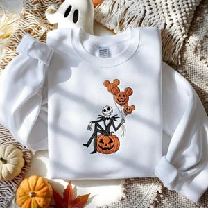 May include: A white crewneck sweatshirt featuring an embroidered graphic of a skeleton character in a pinstripe suit sitting on a large orange jack-o'-lantern. The character holds three orange pumpkin balloons shaped like mouse ears.