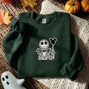 May include: A dark green crewneck sweatshirt featuring a white embroidered graphic of a skeletal character in a pinstriped suit holding a black balloon shaped like a mouse head. The garment has long sleeves, a ribbed neckline, and a white brand label.