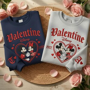 Puede incluir: Dos sudaderas, una azul marino y otra gris, con un dise&ntilde;o de San Valent&iacute;n. Cada sudadera tiene la palabra "Valentine" en escritura roja, el logotipo de Disney y un coraz&oacute;n con Mickey y Minnie Mouse. Las mangas tienen las letras "T" y "A".