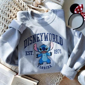 May include: A gray sweatshirt with the words "DISNEYWORLD" embroidered in blue, along with a blue cartoon character and the text "EST. 1971 FLORIDA." A black headband with Minnie Mouse ears and a red bow is also visible.