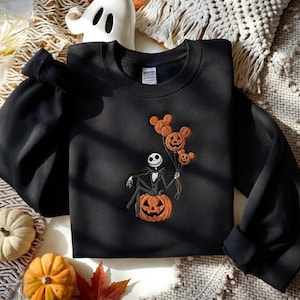 May include: A black crewneck sweatshirt featuring an embroidered design of a skeleton character sitting on a large orange jack-o'-lantern. The character holds three orange pumpkin balloons shaped like mouse ears with carved faces.
