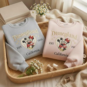May include: Two sweatshirts, one gray and one pink, with the Disneyland logo embroidered in gold. The logo features Mickey and Minnie Mouse in a heart, with "EST 1955 California" below. The sweatshirts are in a woven tray.