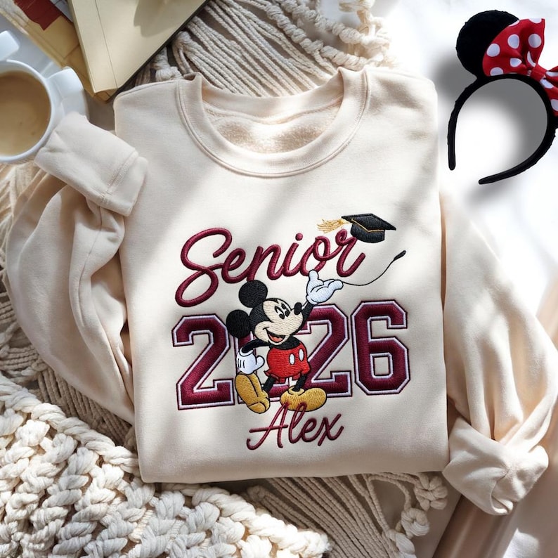 May include: Cream-colored sweatshirt with embroidered "Senior 2026" text and a Mickey Mouse graphic. The design includes a graduation cap and the name "Alex" below the numbers. A Mickey Mouse ear headband with a red bow is also visible.