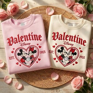 Puede incluir: Dos sudaderas, una rosa y otra crema, con un dise&ntilde;o de San Valent&iacute;n. Cada sudadera tiene la palabra "Valentine" en escritura roja, el logo de Disney y un coraz&oacute;n con Mickey y Minnie Mouse. P&eacute;talos de rosa rosa rodean las sudaderas.