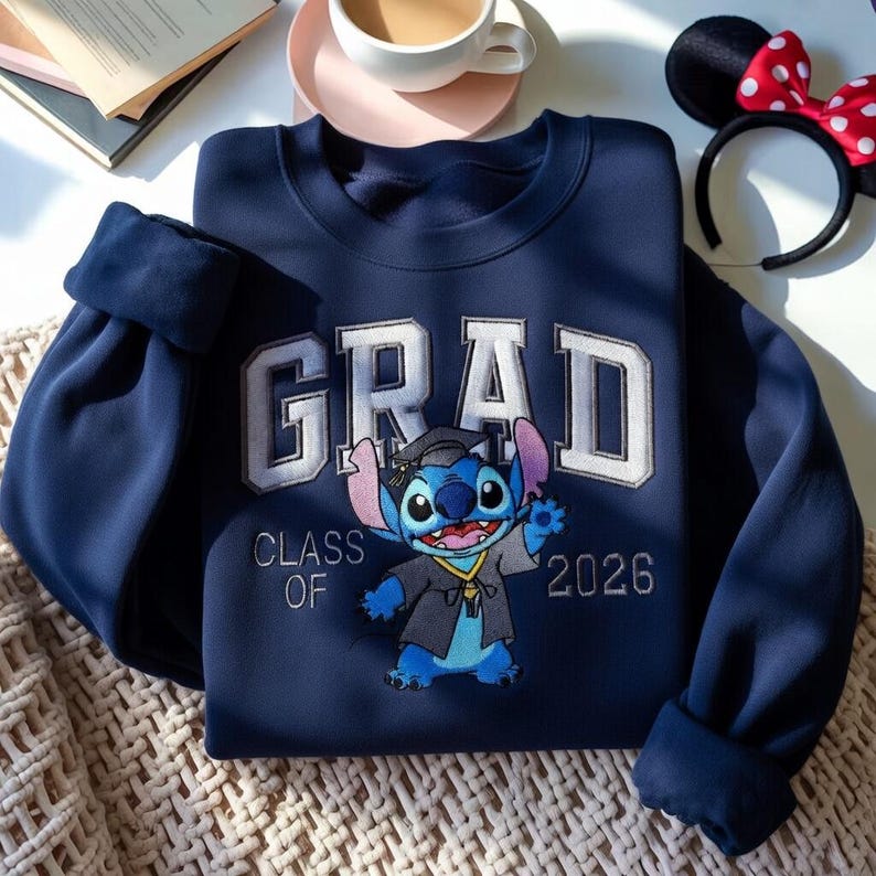 Puede incluir: Sudadera azul marino con la palabra "GRAD" en letras blancas y un personaje de Stitch con un atuendo de graduaci&oacute;n. Tambi&eacute;n se muestra el texto "CLASS OF 2026". La sudadera tiene mangas con pu&ntilde;os.