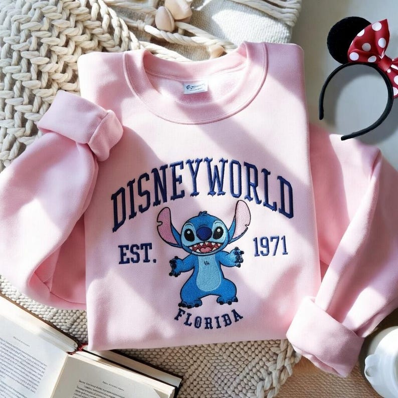 May include: A light pink sweatshirt featuring the text "DISNEYWORLD" above a blue cartoon character, with "EST. 1971 FLORIDA" below. A black headband with Minnie Mouse ears and a red polka dot bow is visible.