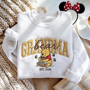 May include: White sweatshirt with the word "Grandma" in gold lettering, featuring a Winnie the Pooh graphic holding a honey pot. The sweatshirt also has the text "EST. 2026" below the graphic. A Minnie Mouse headband is visible.