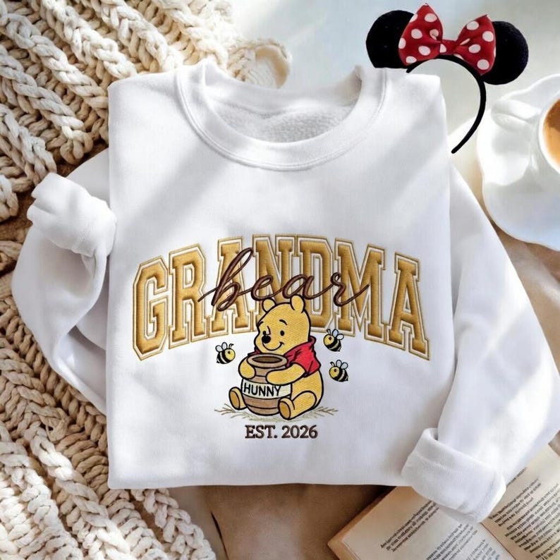 May include: White sweatshirt with the word "Grandma" in gold lettering, featuring a Winnie the Pooh graphic holding a honey pot. The sweatshirt also has the text "EST. 2026" below the graphic. A Minnie Mouse headband is visible.