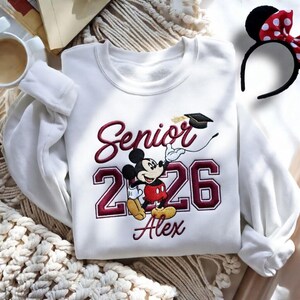 May include: White sweatshirt with embroidered design featuring Mickey Mouse in graduation attire. The design includes the word "Senior" and the year "2026" in maroon, along with the name "Alex". A Minnie Mouse ear headband with a red bow is also visible.