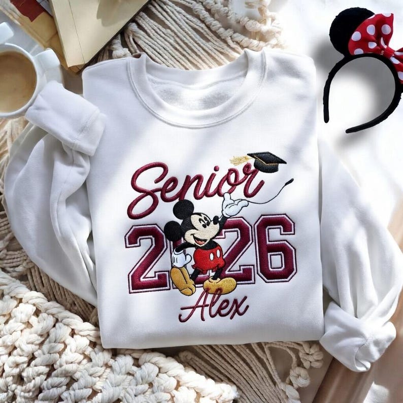 May include: White sweatshirt with embroidered design featuring Mickey Mouse in graduation attire. The design includes the word "Senior" and the year "2026" in maroon, along with the name "Alex". A Minnie Mouse ear headband with a red bow is also visible.