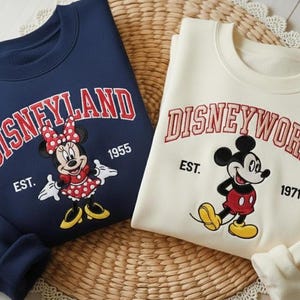 May include: Two sweatshirts, one navy blue and one cream-colored, featuring Disney characters. The navy sweatshirt says "Disneyland" with Minnie Mouse, and the cream one says "Disneyworld" with Mickey Mouse.