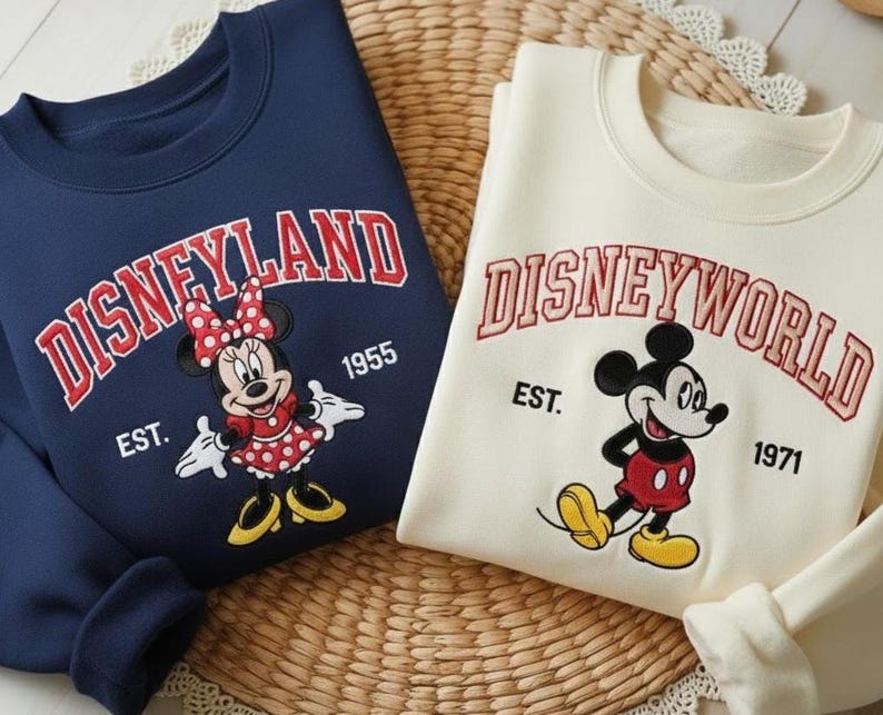 May include: Two sweatshirts, one navy blue and one cream-colored, featuring Disney characters. The navy sweatshirt says "Disneyland" with Minnie Mouse, and the cream one says "Disneyworld" with Mickey Mouse.