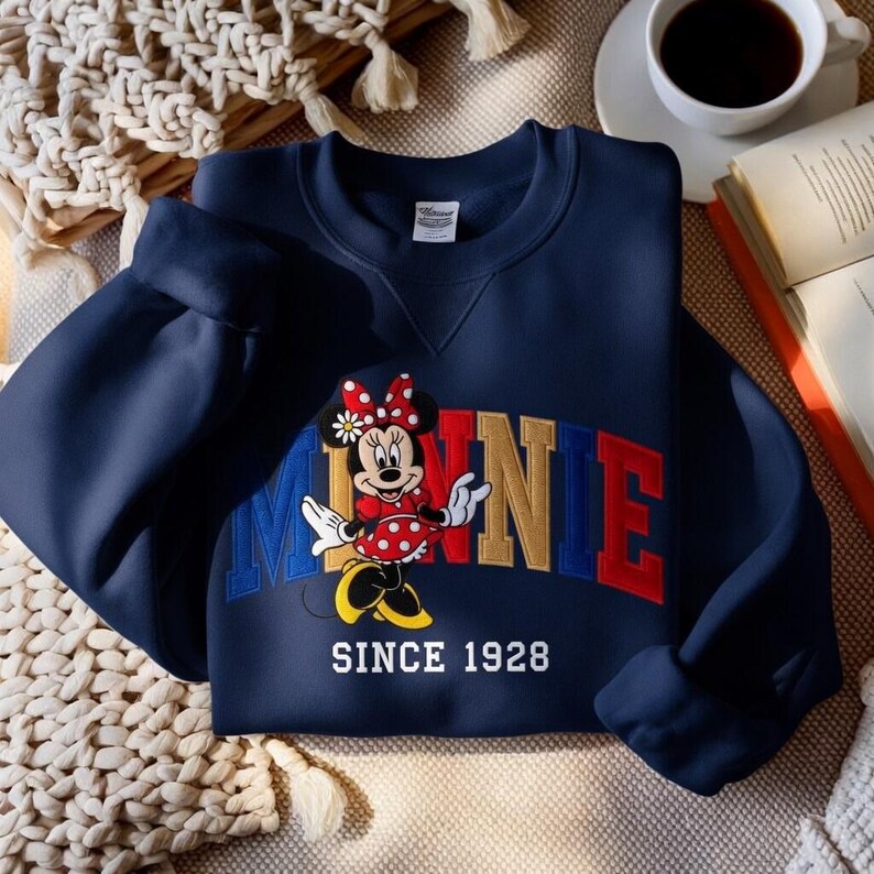K&ouml;nnte beinhalten: Marineblaues Sweatshirt mit Minnie Mouse-Motiv. Der Schriftzug "MINNIE" ist in Rot, Gold und Blau gehalten, mit Minnie Mouse in einem roten gepunkteten Kleid. Der Text "SINCE 1928" ist darunter gedruckt.