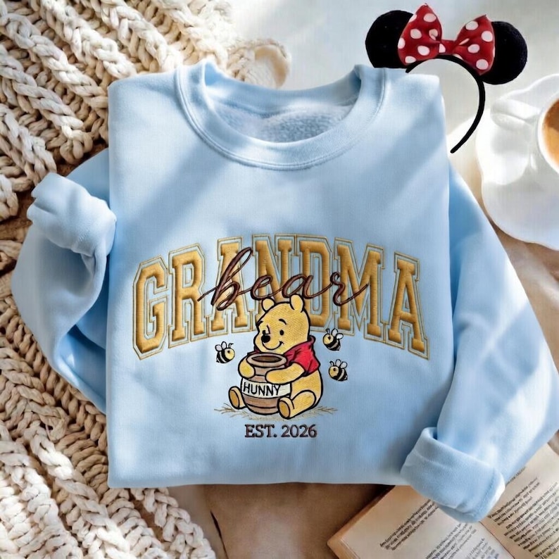 May include: Light blue sweatshirt with "Grandma Bear" embroidered in gold lettering, featuring Winnie the Pooh holding a honey pot. Includes "EST. 2026" and three bees. A Minnie Mouse ear headband is visible.