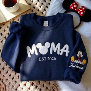 May include: Navy blue sweatshirt with the word "MOM" in white, featuring a Mickey Mouse head for the "O". The sleeve has a Mickey Mouse patch and the name "Jackson". The text "EST.2026" is below the word "MOM".
