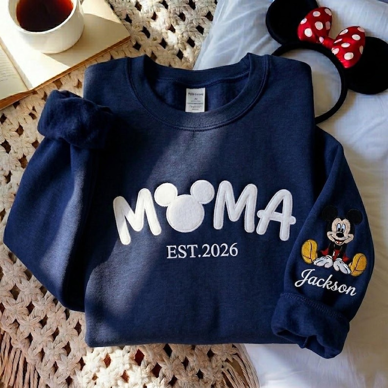 May include: Navy blue sweatshirt with the word "MOM" in white, featuring a Mickey Mouse head for the "O". The sleeve has a Mickey Mouse patch and the name "Jackson". The text "EST.2026" is below the word "MOM".
