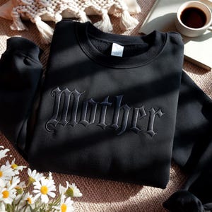 Könnte beinhalten: Schwarzes Sweatshirt mit Rundhalsausschnitt und dem Wort "Mother" in gotischer Schrift gestickt. Das Sweatshirt besteht aus einem weichen Material. Das Design ist auf der Brust zentriert. Die Farbe ist dunkel.