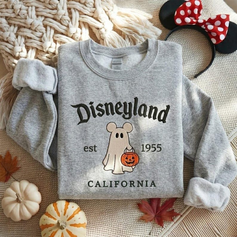 Pode incluir: Uma camisola cinzenta mesclada de gola redonda com texto bordado a preto que diz Disneyland California. Abaixo do texto, um fantasma bordado com orelhas de rato segura uma ab&oacute;bora laranja, com o texto est 1955 nos lados.